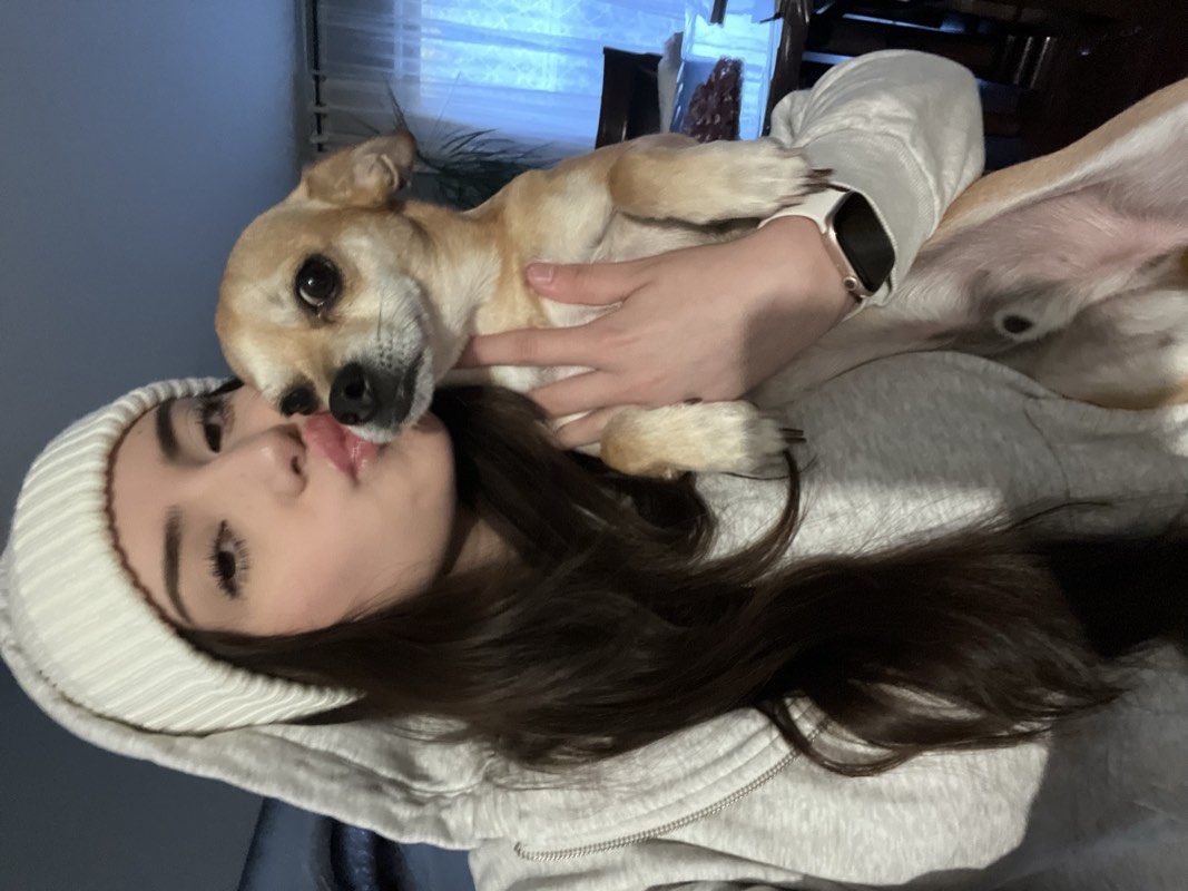 Woman in a cream knit hat and gray hoodie holding a small tan dog close to her face.